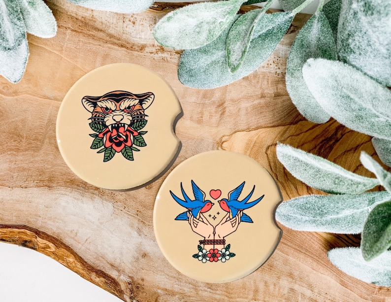 May include: Two round beige coasters with colorful illustrations. One coaster features a tiger with a red rose in its mouth. The other coaster features two blue birds with a red heart between them.