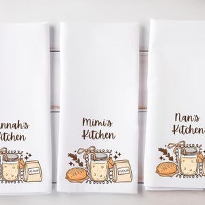 May include: Three white kitchen towels with different text: "Hannah's Kitchen", "Mimi's Kitchen", and "Nan's Kitchen". Each towel has a similar illustration of a jar, bread, and a bag of flour. The illustrations are in brown, beige, and tan.