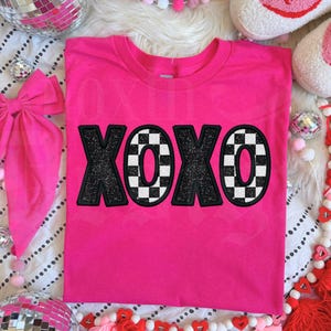 May include: A bright pink t-shirt with the letters "XOXO" in a black glitter and checkered design. The shirt is surrounded by decorative elements, including a large pink bow, disco balls, and pom-poms.