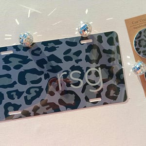 May include: A black and gray leopard print car license plate with the letters "rsg" in white. The license plate is next to two gray and black leopard print car coasters with the text "Car Coasters Handmade with love" on a white card.