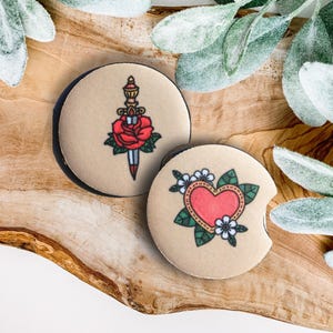 May include: Two round beige coasters with black trim. One coaster features a red rose with a dagger through it. The other coaster features a red heart with green leaves and white flowers.