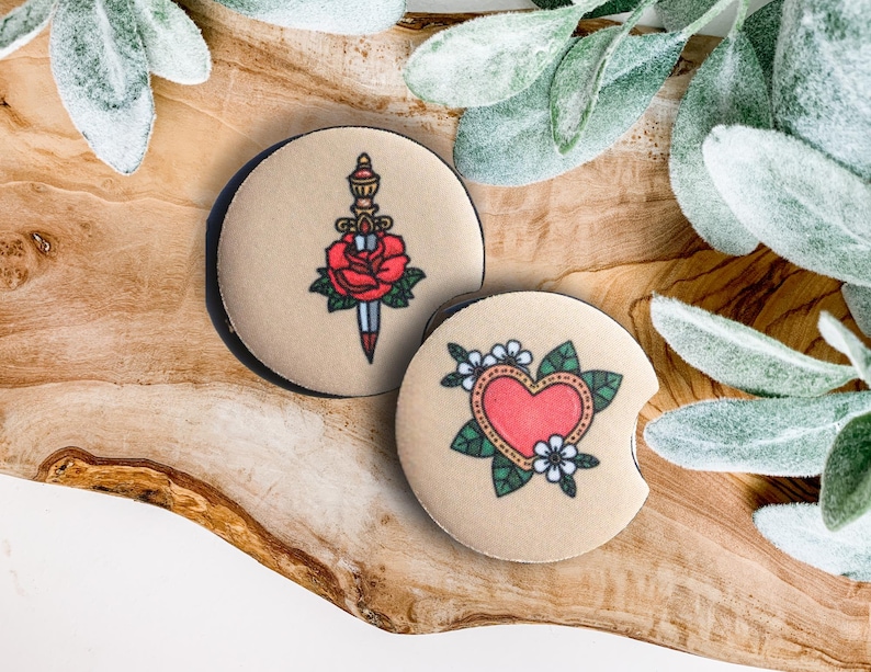 May include: Two round beige coasters with black trim. One coaster features a red rose with a dagger through it. The other coaster features a red heart with green leaves and white flowers.