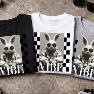 May include: Three t-shirts in black, white, and gray, each featuring a graphic of a rabbit wearing sunglasses and a jacket. The word "VIBE" is printed below the rabbit image. A black and white checkered pattern frames the rabbit image.