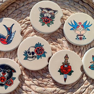 May include: Set of six round coasters with various vintage tattoo-inspired designs. The coasters feature a panther, a dagger with a rose, a skull with a rose, a swallow, two birds, and a tiger. The coasters are beige with black outlines.