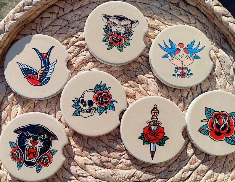May include: Set of six round coasters with various vintage tattoo-inspired designs. The coasters feature a panther, a dagger with a rose, a skull with a rose, a swallow, two birds, and a tiger. The coasters are beige with black outlines.