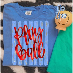 May include: A light blue t-shirt with the words "Play Ball" in red script. The shirt has vertical blue and gold stripes. A small, smiling, yellow ceramic figure and a green shirt are also visible.