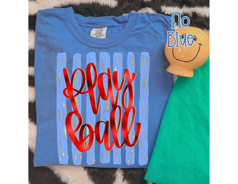 May include: A light blue t-shirt with the words "Play Ball" in red script. The shirt has vertical blue and gold stripes. A small, smiling, yellow ceramic figure and a green shirt are also visible.