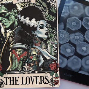 May include: A tarot card featuring a gothic illustration of Frankenstein's monster and his bride, with the text "THE LOVERS". The card has a vintage aesthetic with a black, green, and red color scheme. A Kindle e-reader is visible in the background.