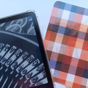 May include: A digital tablet in a clear case shows a monochrome image of typewriter keys. Next to it is a card with a plaid pattern in orange, red, blue, and white.