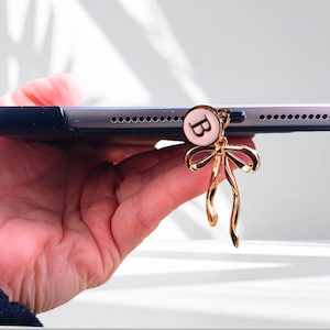 May include: A gold-colored bow charm with a small, round, pink and white pendant featuring the letter "B" is attached to a tablet. The charm is hanging from the side of the tablet.