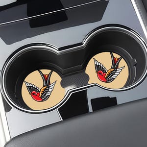 May include: Two beige car coasters with a vintage-style swallow tattoo design. The swallow is red, black, and yellow.