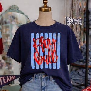 May include: Navy blue t-shirt with the text "Play Ball" in red script, set against a background of blue vertical stripes. The shirt is displayed on a mannequin.
