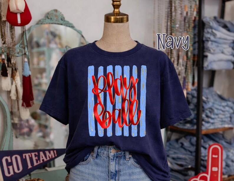May include: Navy blue t-shirt with the text "Play Ball" in red script, set against a background of blue vertical stripes. The shirt is displayed on a mannequin.