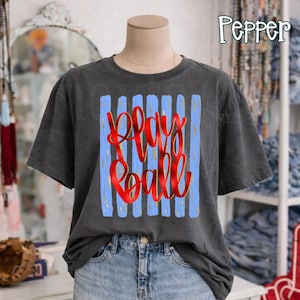 May include: A charcoal gray t-shirt with a graphic that says "Play Ball" in red script over blue vertical stripes. The shirt is displayed on a mannequin, paired with denim shorts. The word "Pepper" is in the top right corner.