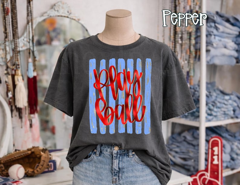 May include: A charcoal gray t-shirt with a graphic that says "Play Ball" in red script over blue vertical stripes. The shirt is displayed on a mannequin, paired with denim shorts. The word "Pepper" is in the top right corner.