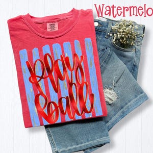 May include: A coral-colored t-shirt with the words "Play Ball" in red script over blue vertical stripes. The shirt is paired with denim shorts and a sprig of white flowers.
