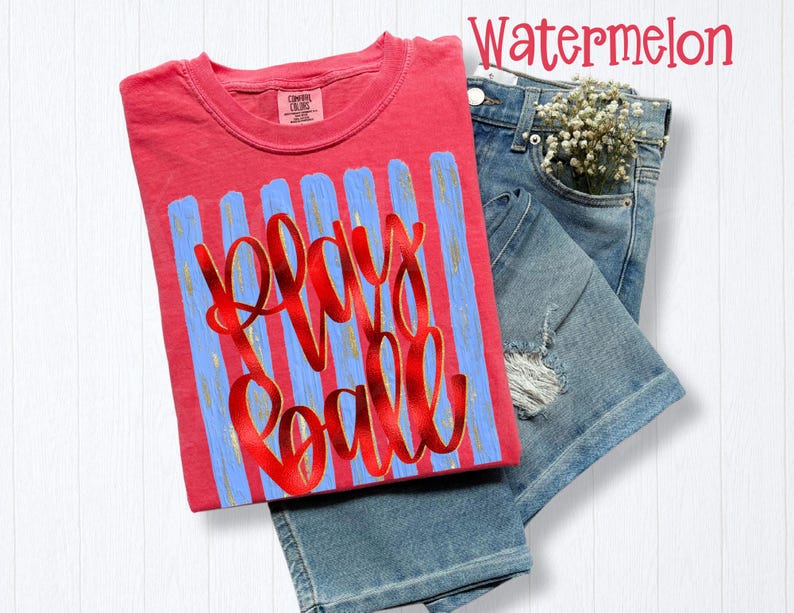 May include: A coral-colored t-shirt with the words "Play Ball" in red script over blue vertical stripes. The shirt is paired with denim shorts and a sprig of white flowers.