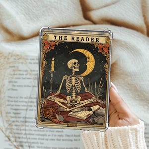 May include: A clear phone case featuring a tarot card design. The card depicts a skeleton seated in a meditative pose, with a crescent moon, candle, and the text "THE READER". The card has a dark background with gold and red accents.