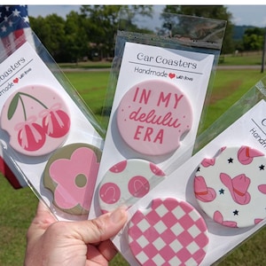 May include: Set of six pink and white car coasters with various designs. The coasters feature a cherry, a flower, polka dots, a checkered pattern, and a cowboy hat. The coasters are packaged in clear plastic bags with a white label that reads "Car Coasters Handmade with love".