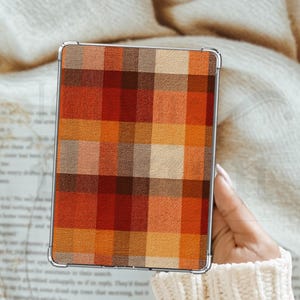 May include: A rectangular device case featuring a plaid design in autumnal shades of orange, red, and brown. The case has a silver frame and is held against a cream-coloured, textured background.