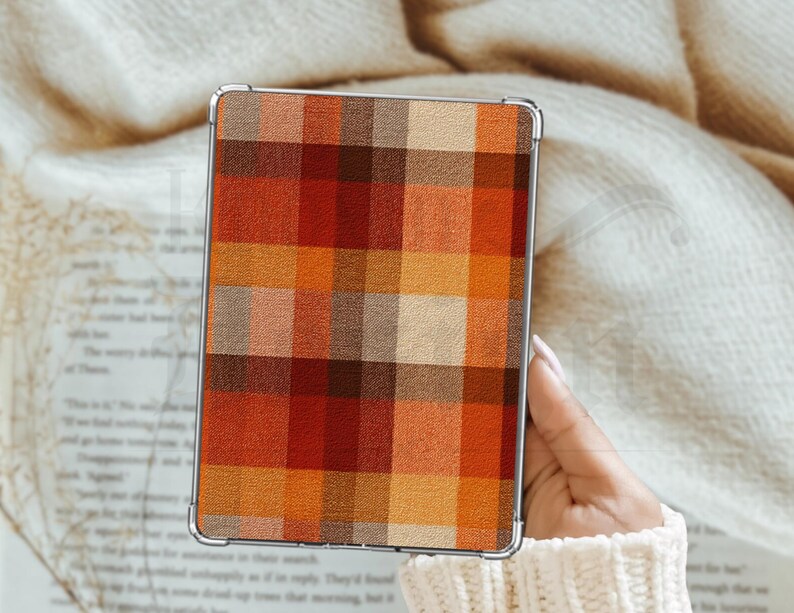 May include: A rectangular device case featuring a plaid design in autumnal shades of orange, red, and brown. The case has a silver frame and is held against a cream-coloured, textured background.