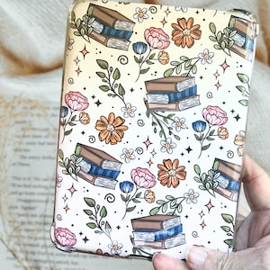 May include: A cream-colored case with a repeating pattern of books, flowers, and leaves. The books are brown with blue pages. The flowers are pink, orange, and white. The case is held in a hand.