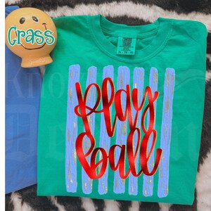 May include: A green t-shirt with the words "Play Ball" in red script. The shirt has vertical blue stripes with gold accents. A small, smiling, yellow figurine with the word "Crass" is in the upper left corner.