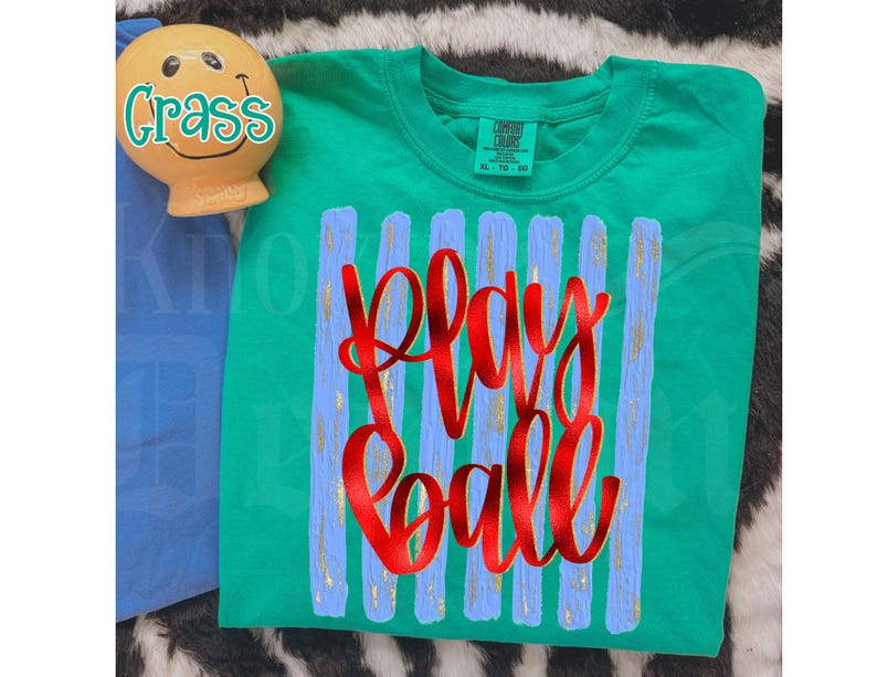 May include: A green t-shirt with the words "Play Ball" in red script. The shirt has vertical blue stripes with gold accents. A small, smiling, yellow figurine with the word "Crass" is in the upper left corner.