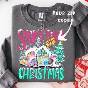 May include: Dark gray sweatshirt with a colorful "Small Town Christmas" graphic. The design features snow-covered houses, a snowman, and a Christmas tree. The text "Your Zip Code" is also visible.