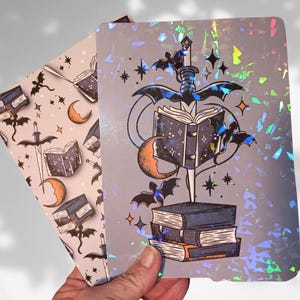 May include: Two decorative notebooks with a fantasy theme. One notebook features a repeating pattern of bats, books, swords, and moons. The other notebook has a design of a sword piercing a stack of books, with bats, stars, and a crescent moon.