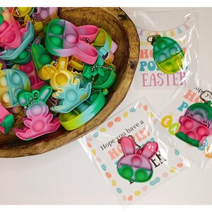 May include: A bowl filled with colorful Easter-themed pop it fidget toys shaped like bunnies and eggs. The toys are attached to keychains and have a rainbow swirl design. The text on the card reads "Hope you have a Hoppy Easter!"