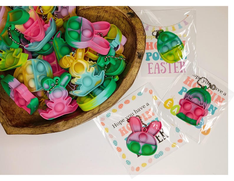 May include: A bowl filled with colorful Easter-themed pop it fidget toys shaped like bunnies and eggs. The toys are attached to keychains and have a rainbow swirl design. The text on the card reads "Hope you have a Hoppy Easter!"