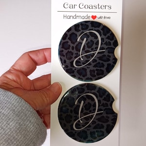 May include: Two black and gray leopard print car coasters with a silver letter D on each. The coasters are handmade with love.