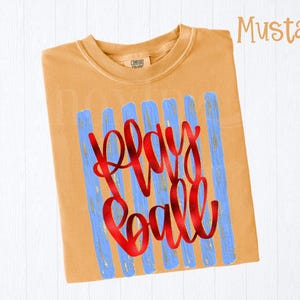 May include: Mustard-colored t-shirt with the words "Play Ball" in red script. The text is superimposed over blue vertical brushstrokes. The shirt has a crew neck and short sleeves.
