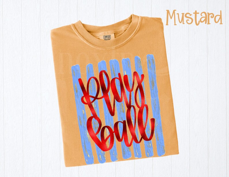 May include: Mustard-colored t-shirt with the words "Play Ball" in red script. The text is superimposed over blue vertical brushstrokes. The shirt has a crew neck and short sleeves.