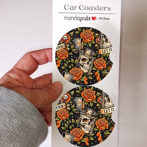 May include: Two car coasters with a black background featuring a skull, roses, and a lighter design. The coasters have the words "BURN" and "LET IT" on them. The coasters are held by a person.