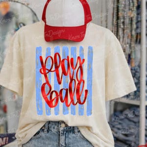 May include: Cream-colored t-shirt with blue vertical stripes and the words "Play Ball" in red script. A red and white baseball cap rests on top of the shirt. The shirt is made of a soft material.