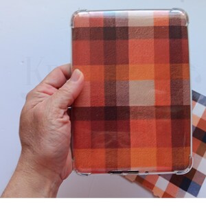 May include: A clear protective case featuring a plaid design in orange, red, and brown hues. The case is held in a hand, with a matching plaid card visible. The case is designed for a tablet or e-reader.