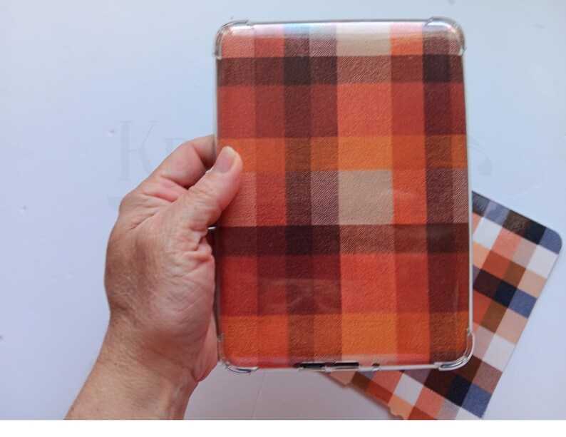 May include: A clear protective case featuring a plaid design in orange, red, and brown hues. The case is held in a hand, with a matching plaid card visible. The case is designed for a tablet or e-reader.