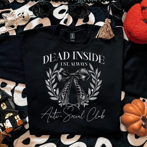 May include: Black sweatshirt with the text "DEAD INSIDE EST. ALWAYS Anti-Social Club" in white script. The design includes a bow and laurel wreath. Halloween-themed decorations surround the sweatshirt, including pumpkins and a skeleton.