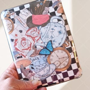 May include: A decorative phone case featuring an Alice in Wonderland theme. The design includes a Mad Hatter hat, playing cards, roses, teacups, a blue butterfly, and pocket watches. The background has a black and white checkered pattern with the text "Eat Me" and "Drink Me".