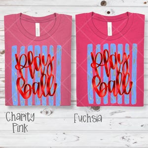 May include: Two pink t-shirts with the words "Play Ball" in red script over blue vertical stripes. The shirt on the left is "Charity Pink" and the shirt on the right is "Fuchsia". Both shirts have a crew neck.