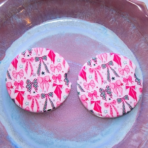 May include: Two pink car coasters with a repeating pattern of bows, stars, and checkered ribbons. The coasters have a small notch for easy removal from a car cup holder. The background is a pink and purple ceramic surface.