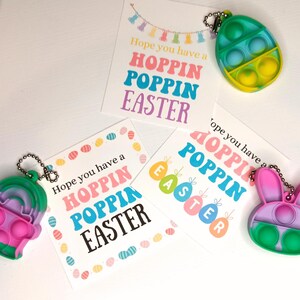 May include: Three colorful Easter-themed pop-it fidget toys with keychains. The toys are shaped like an egg, a bunny, and a cupcake. Each toy has a different color scheme and features a pop-it design. The toys are accompanied by three cards with the text "Hope you have a Hoppin Poppin Easter".