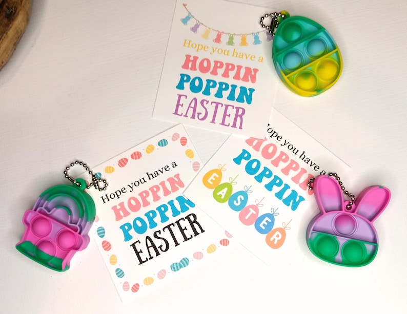 May include: Three colorful Easter-themed pop-it fidget toys with keychains. The toys are shaped like an egg, a bunny, and a cupcake. Each toy has a different color scheme and features a pop-it design. The toys are accompanied by three cards with the text "Hope you have a Hoppin Poppin Easter".