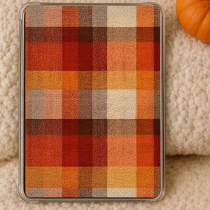 May include: A tablet case featuring a plaid design in autumnal shades of orange, red, and brown. The case is set against a cream-coloured, textured backdrop, with a small orange pumpkin visible.