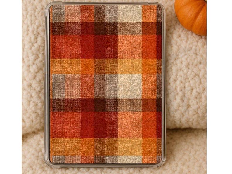 May include: A tablet case featuring a plaid design in autumnal shades of orange, red, and brown. The case is set against a cream-coloured, textured backdrop, with a small orange pumpkin visible.