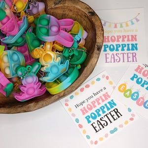 May include: A wooden bowl filled with colorful Easter-themed pop-it fidget toys. The toys are shaped like bunnies, eggs, and other Easter symbols. The bowl is sitting on a white surface next to two cards with the text "Hope you have a Hoppin Poppin Easter".