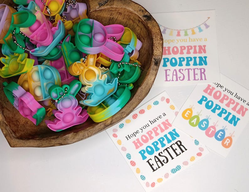 May include: A wooden bowl filled with colorful Easter-themed pop-it fidget toys. The toys are shaped like bunnies, eggs, and other Easter symbols. The bowl is sitting on a white surface next to two cards with the text "Hope you have a Hoppin Poppin Easter".