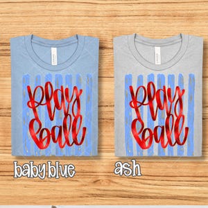 May include: Two t-shirts, one baby blue and one ash, with the words "Play Ball" in red script. The text is superimposed on a blue and gold striped design. The baby blue shirt has the words "baby blue" below it.